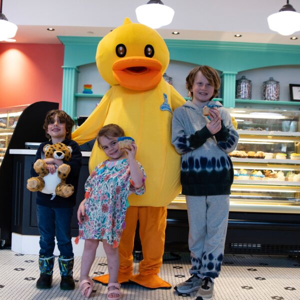 Three smiling kids stand with a person in a duck costume inside a bakery, each holding a treat and stuffed animal, with pastry displays behind them.