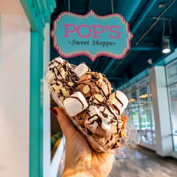 A hand holds a chocolate ice cream sundae with marshmallows under a pink and teal POPS Sweet Shoppe sign inside a brightly lit store.