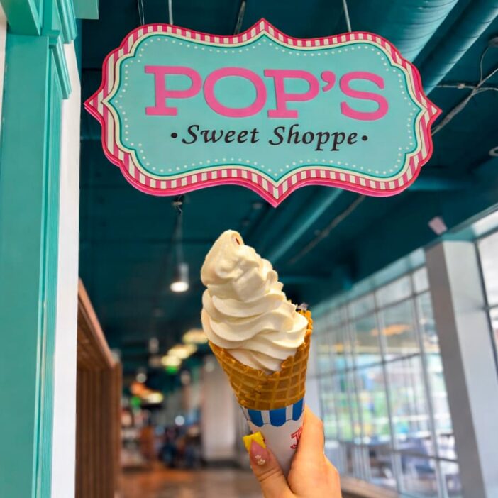 A hand holds a soft serve ice cream cone under a colorful POPS Sweet Shoppe sign inside a shop with teal and pink decor.