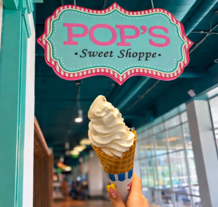 A hand holds a soft serve ice cream cone under a colorful POPS Sweet Shoppe sign inside a shop with teal and pink decor.