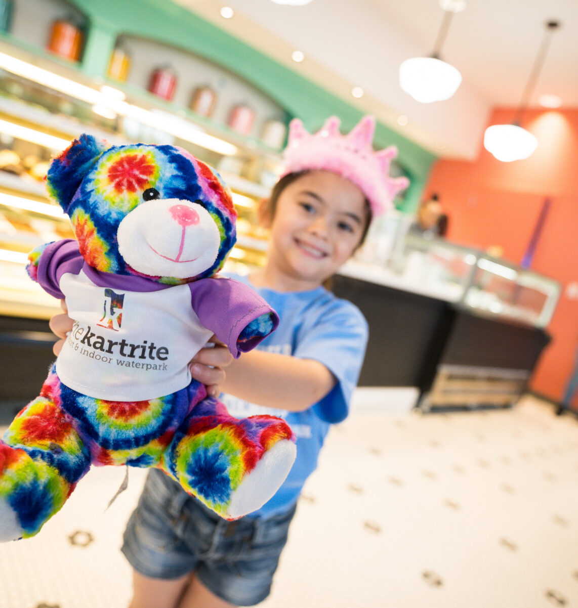 Smiling child in a pink crown holds a colorful tie-dye teddy bear inside a bright bakery or ice cream shop.