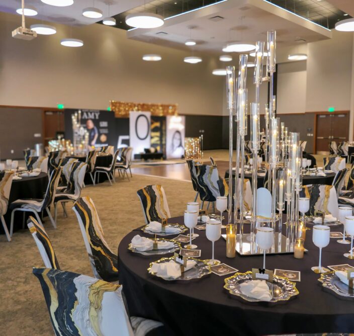 Elegant event hall with round tables set for a celebration, featuring tall candle centerpieces, stylish tableware, and festive decorations in the background.