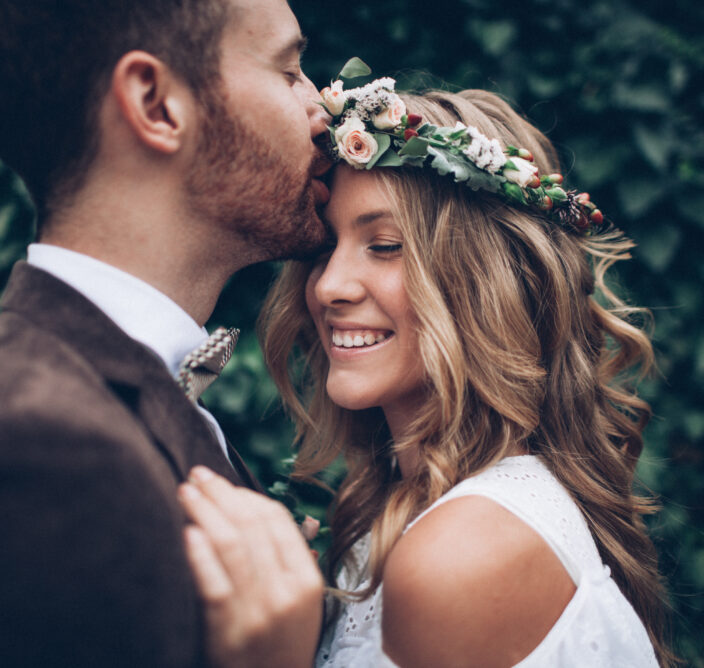 A man kisses a smiling woman on the forehead. She wears a floral crown. Both are dressed up and surrounded by greenery.