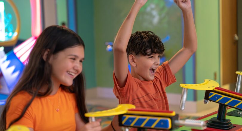 Two kids in orange shirts play an arcade game, smiling and cheering with excitement in a colorful indoor setting.