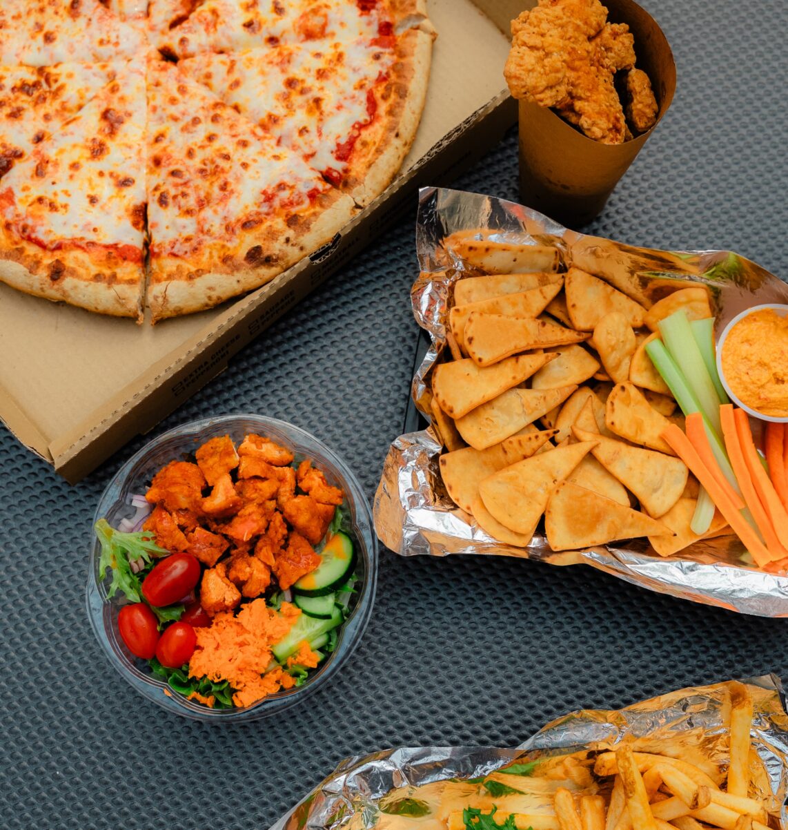 Cheese pizza, fried chicken, salad with buffalo bites, pita chips with dip, veggies, fries, and a sandwich on a textured gray surface.