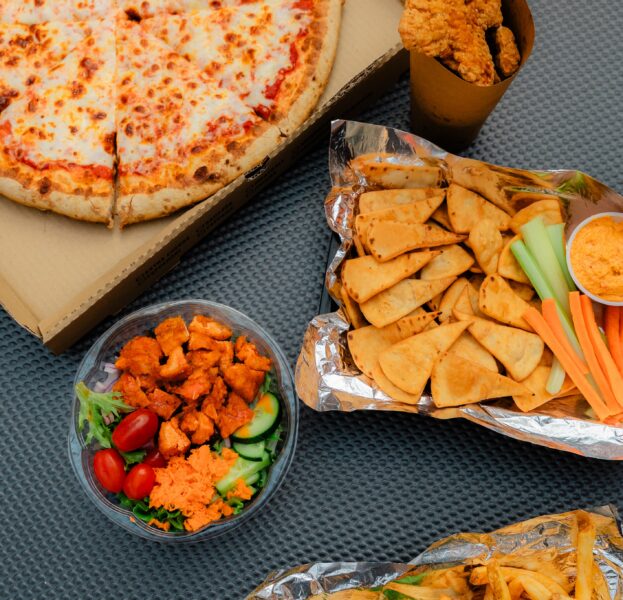 A cheese pizza, fried chicken, pita chips with dip, salad with buffalo chicken, and fries with a sandwich on a gray surface.
