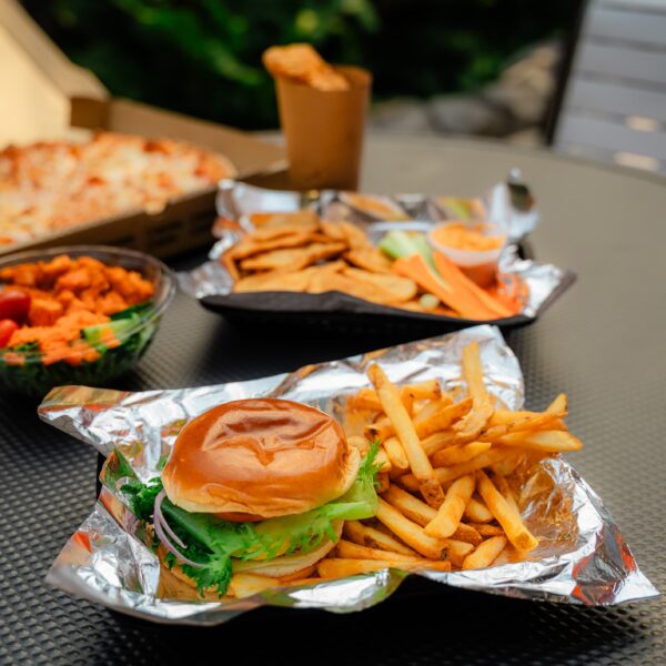Burger and fries on foil, with sides of fries, chicken wings, veggie sticks, and pizza on an outdoor table.