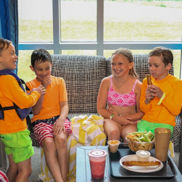 Four kids in swimwear sit on a couch by a window, laughing and eating snacks together. Food and drinks are on a table in front of them.