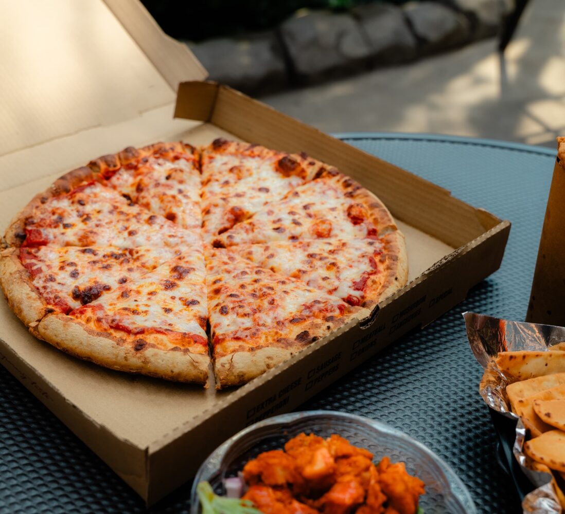 Open pizza box with a cheese pizza, chicken wings, breadsticks, and a cup with a cookie on a table outside.