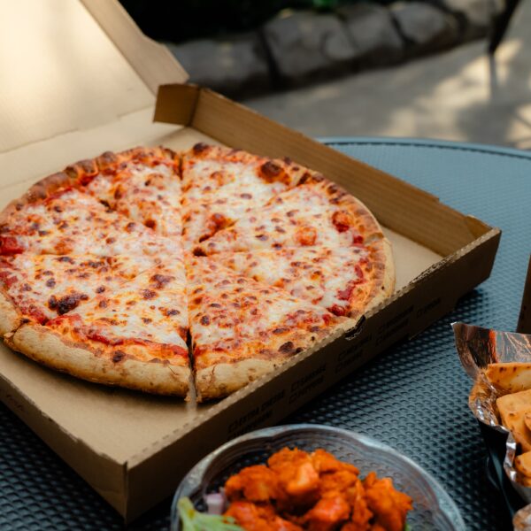 Open pizza box with a cheese pizza, chicken wings, breadsticks, and a cup with a cookie on a table outside.