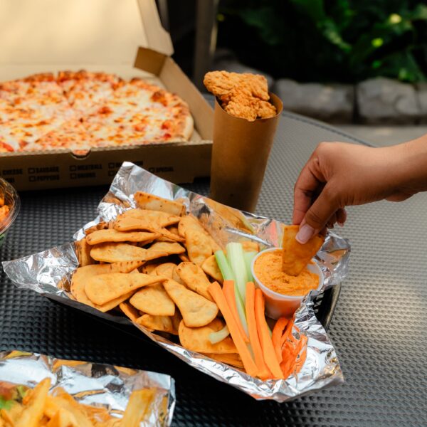 A hand dips a chip in sauce next to carrot and celery sticks, with fries, pizza, and chicken tenders in the background on an outdoor table.