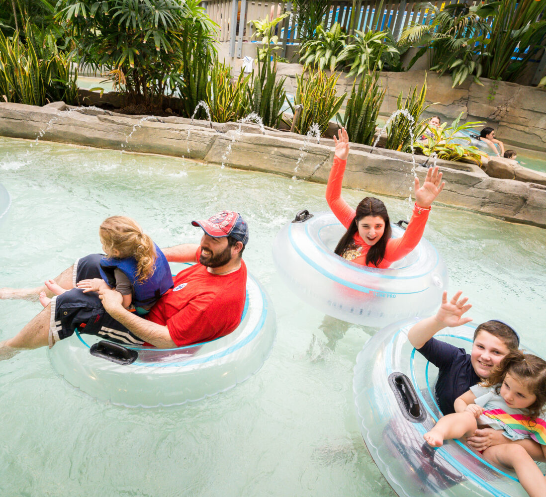 Families float in clear inner tubes on a lazy river, surrounded by lush plants; adults and children smile and wave at the camera.