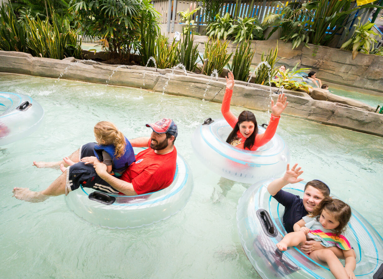 Families float in clear inner tubes on a lazy river, surrounded by lush plants; adults and children smile and wave at the camera.