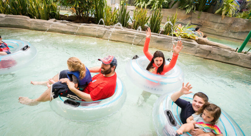 Families float in clear inner tubes on a lazy river, surrounded by lush plants; adults and children smile and wave at the camera.