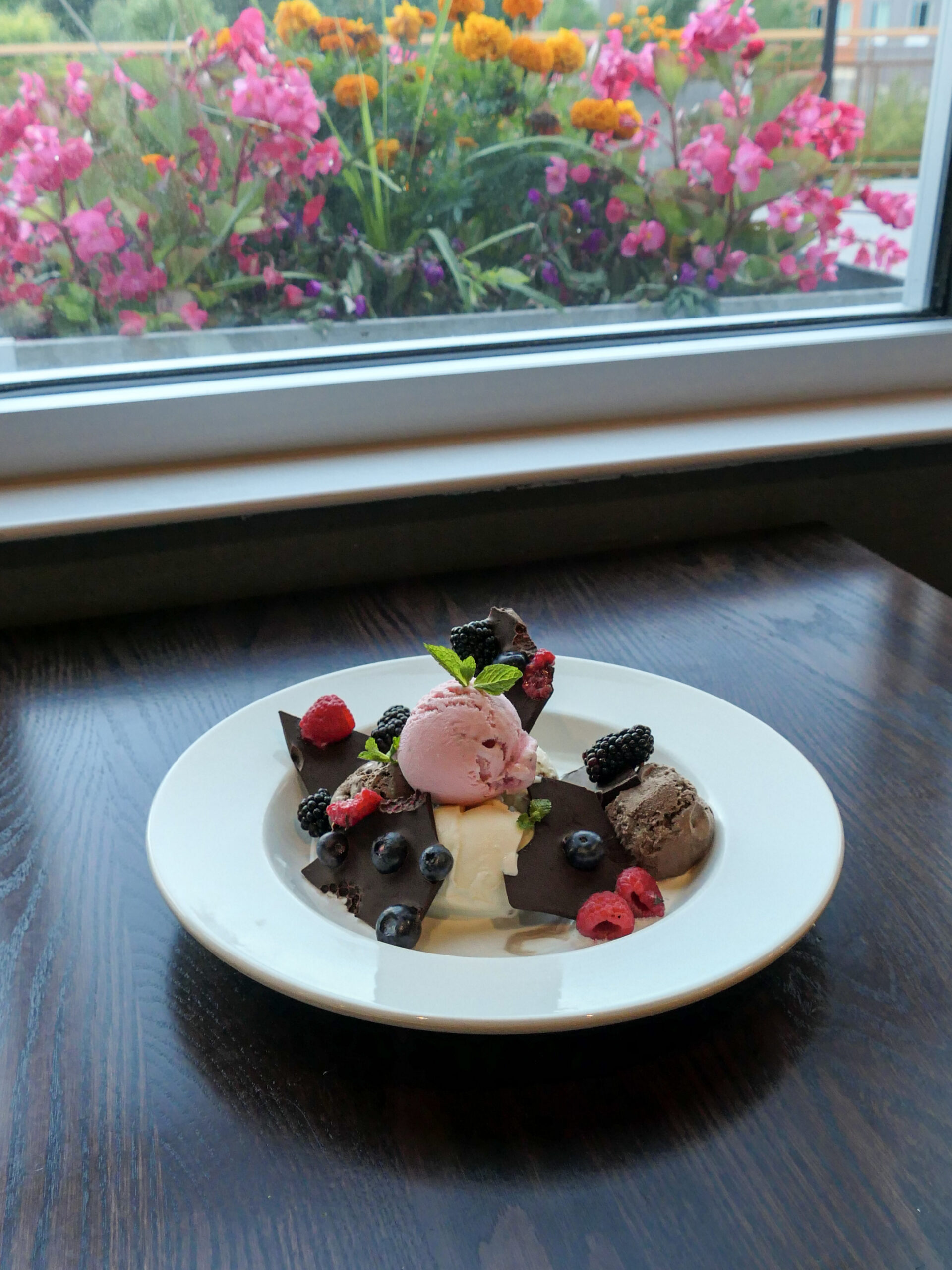 A dessert plate with chocolate and vanilla ice cream, berries, and chocolate pieces sits on a table by a window with colorful flowers outside.