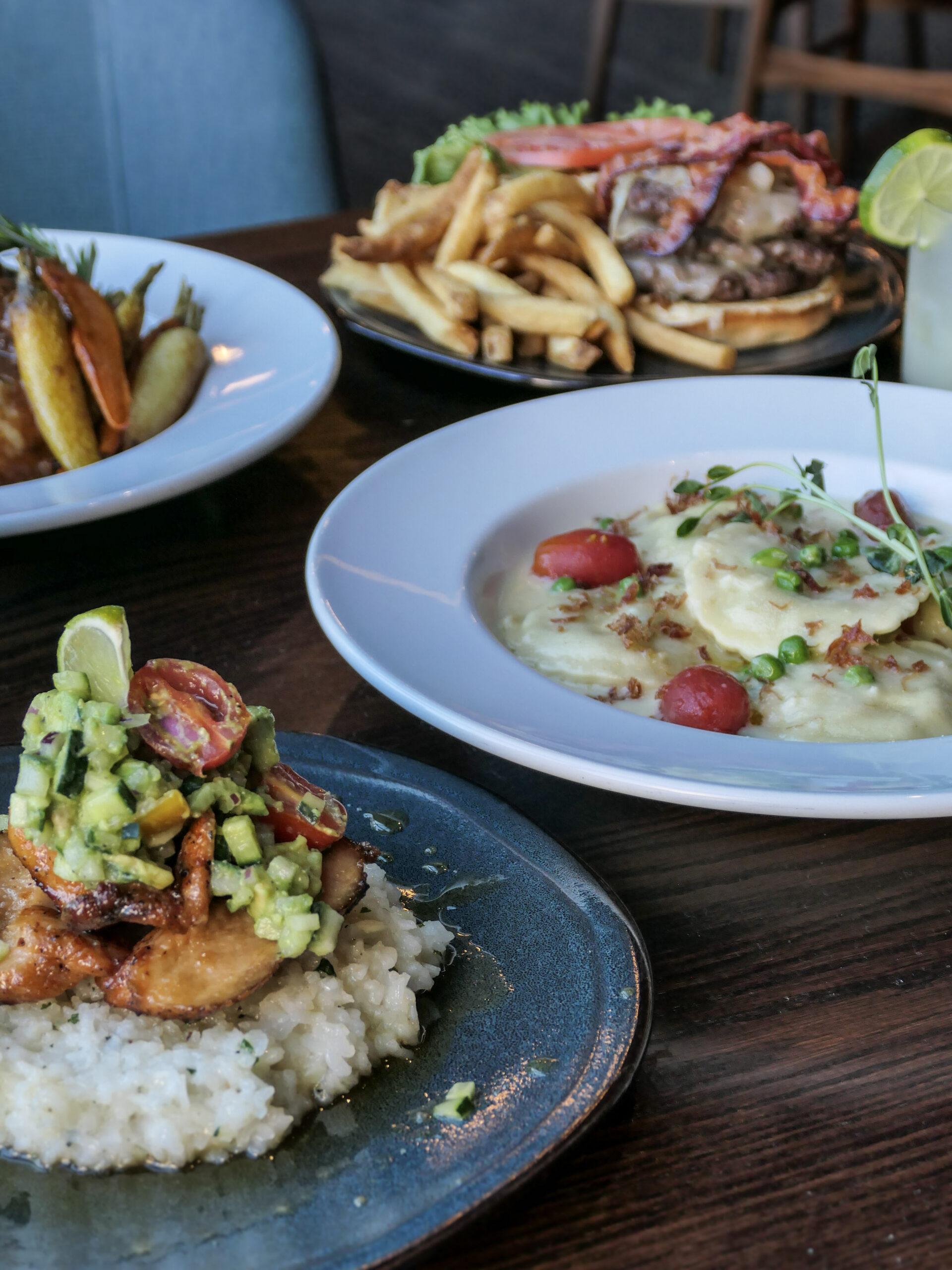 Four plates of food on a wooden table, including fish with rice, mashed potatoes, a burger with fries, and roasted vegetables.