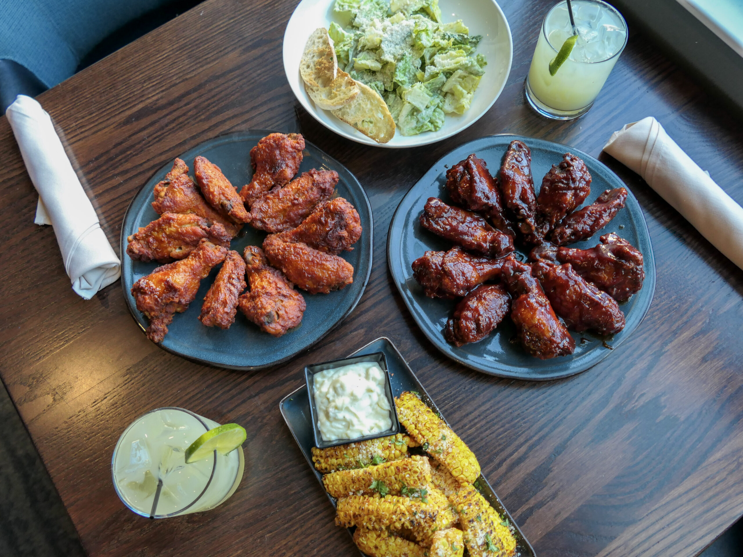 Two plates of chicken wings, a bowl of Caesar salad, corn on the cob, ranch dip, and two cocktails on a wooden table.