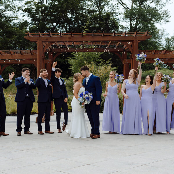 A bride and groom kiss, surrounded by their cheering wedding party in matching blue suits and lavender dresses, outdoors near a wooden gazebo.