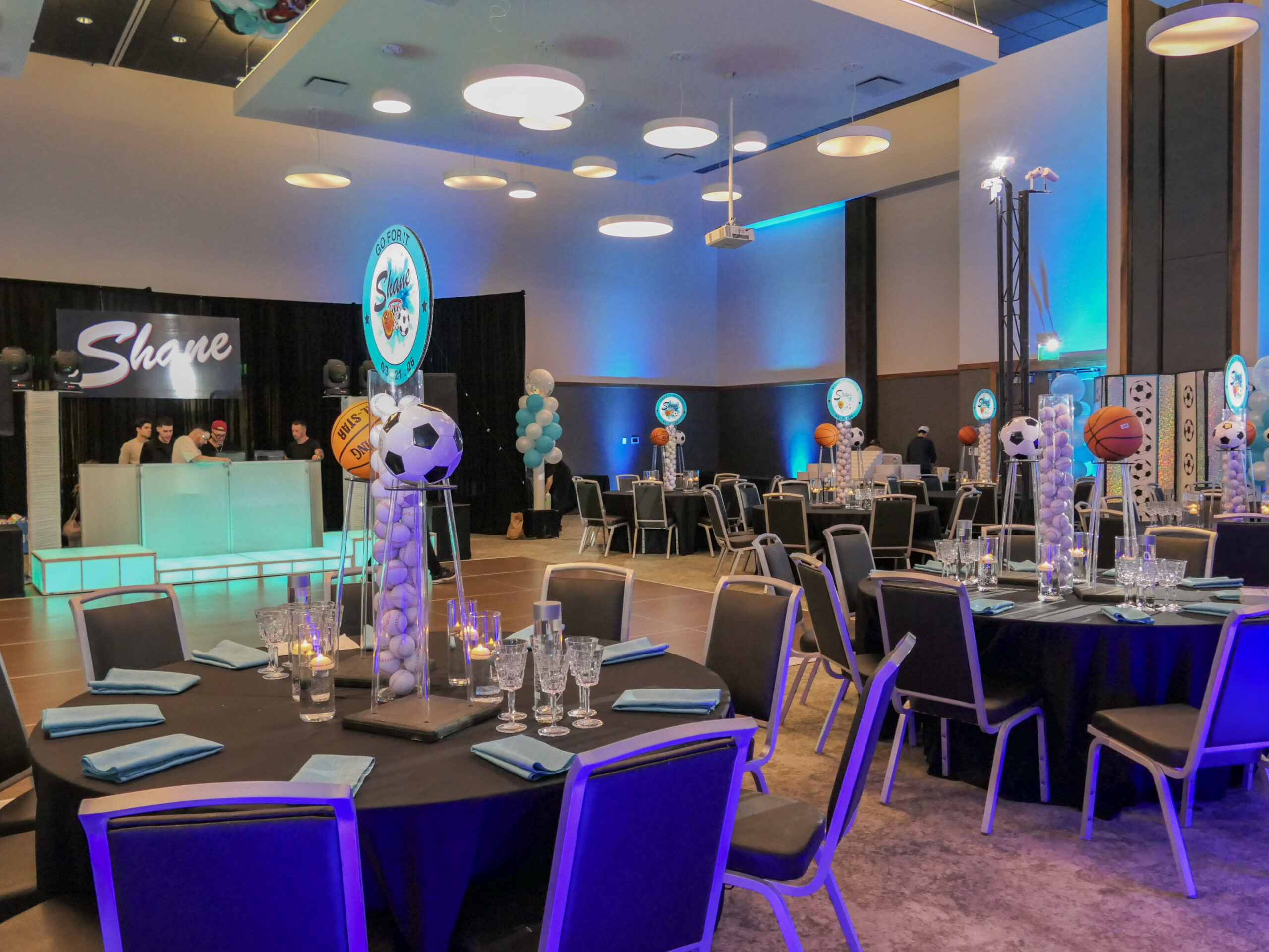 Event hall with sports-themed centerpieces on dining tables, blue lighting, and a DJ booth in the background.
