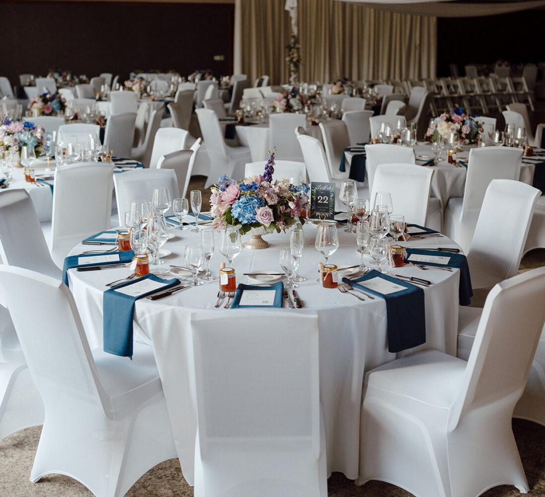 Round tables with white chairs are set for an event, featuring floral centerpieces, glassware, and navy blue napkins in a large, elegant hall.