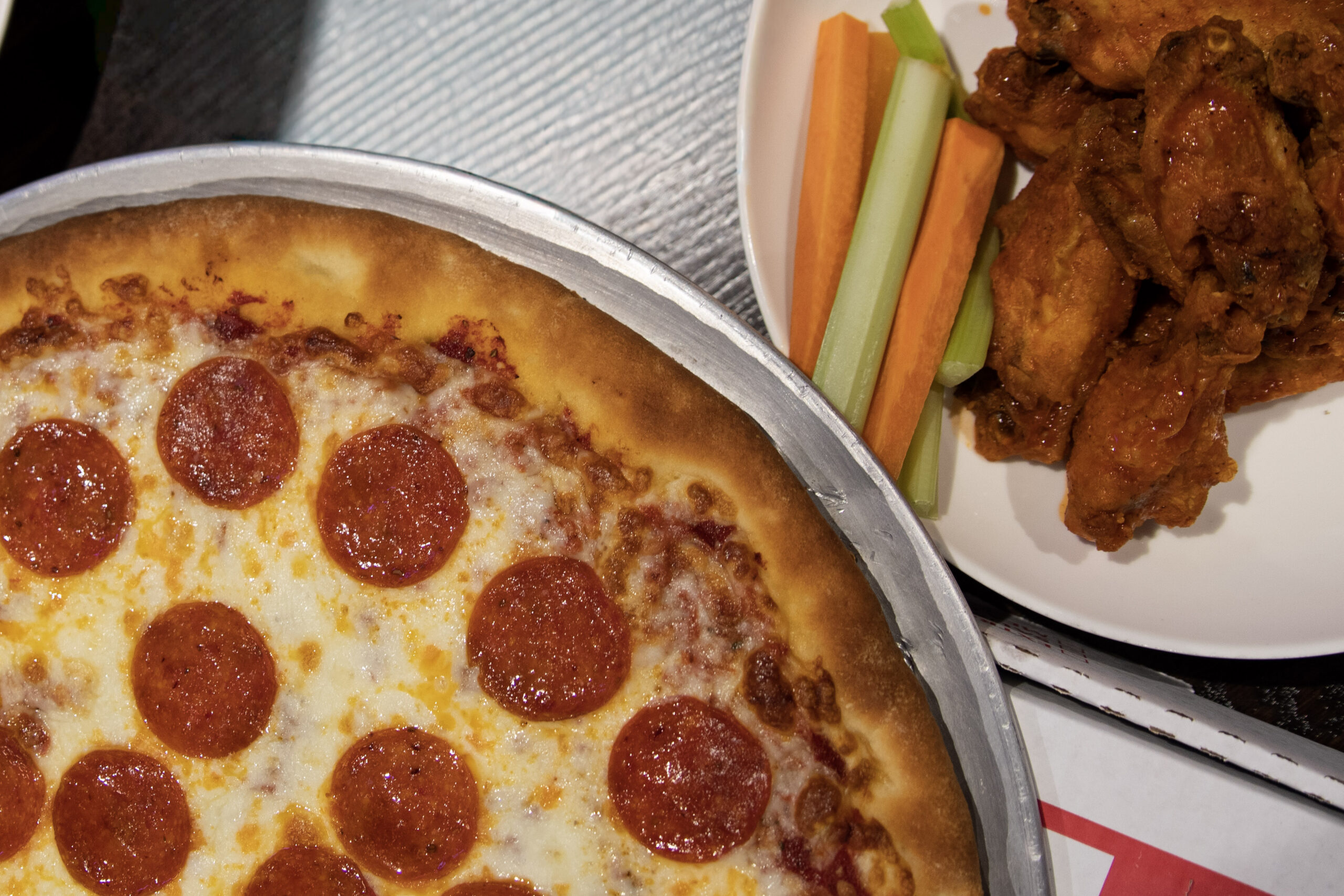 A pepperoni pizza, buffalo chicken wings, and celery and carrot sticks are served on plates on a table.