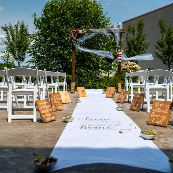 Outdoor wedding aisle with white chairs, a decorated arch, flower petals, and wooden signs with quotes lining the white aisle runner.
