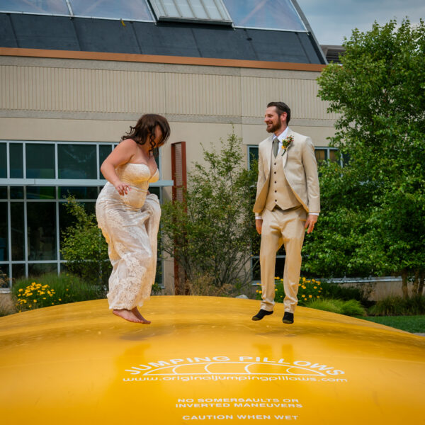 Bride and groom in wedding attire joyfully bounce on a large yellow jumping pillow outdoors, smiling at each other, with greenery and a building behind them.
