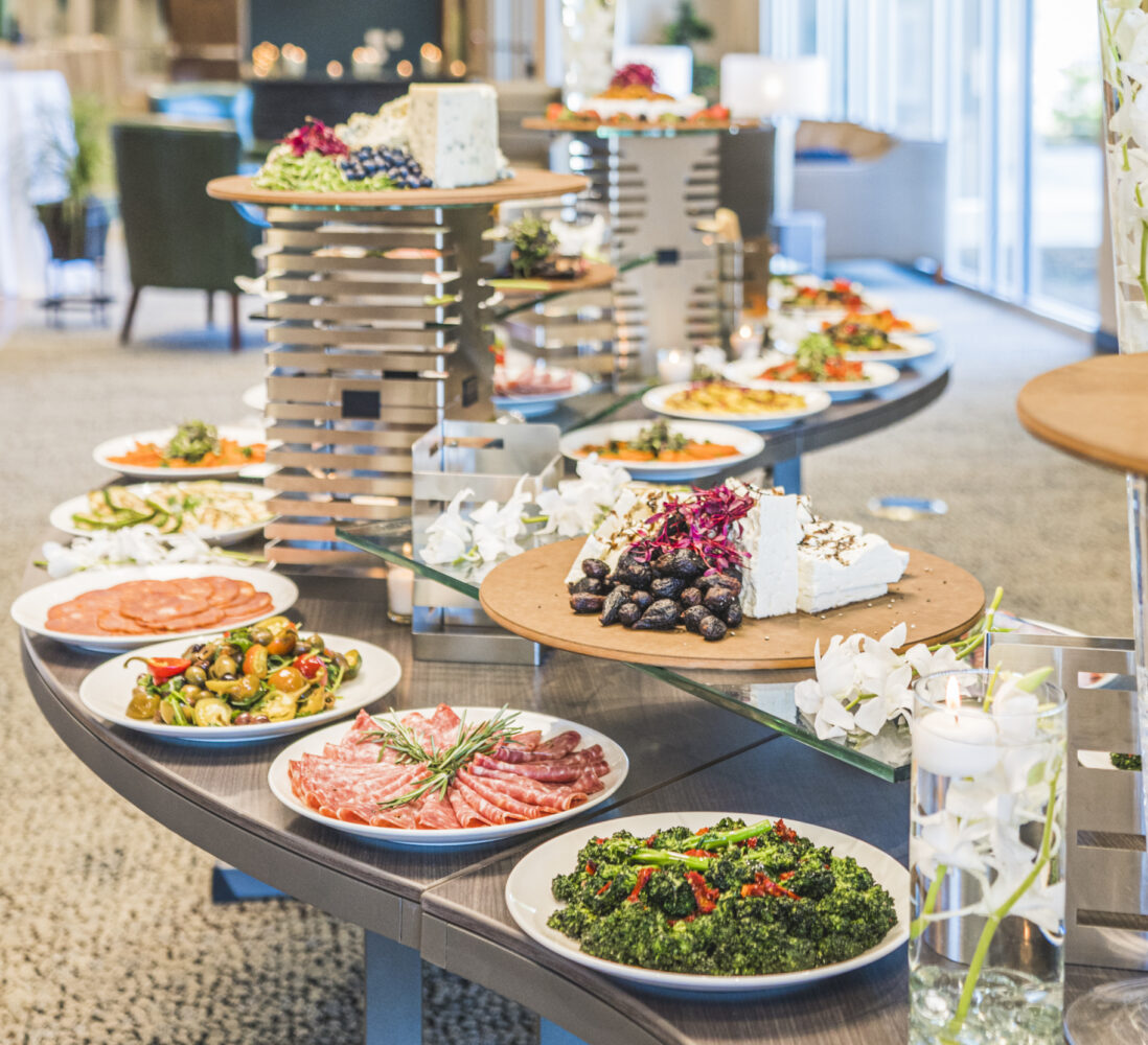 A buffet table displays assorted cheeses, meats, fruits, and salads on white plates in a bright, modern event space.