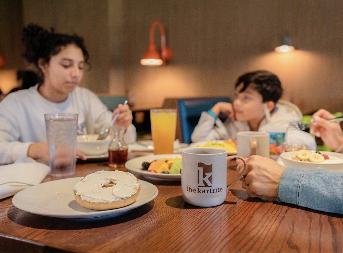 Three people eating breakfast at a restaurant table with a bagel, fruit, drinks, and a the kartrite mug in the foreground.