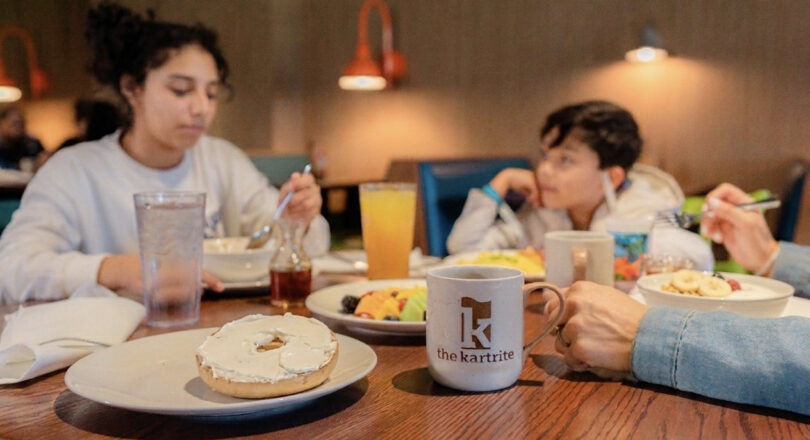 Three people eating breakfast at a restaurant table with a bagel, fruit, drinks, and a the kartrite mug in the foreground.