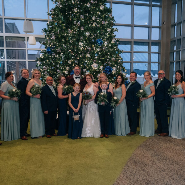 A wedding party poses in front of a large decorated Christmas tree inside a glass-walled venue. Everyone is dressed in formal attire.