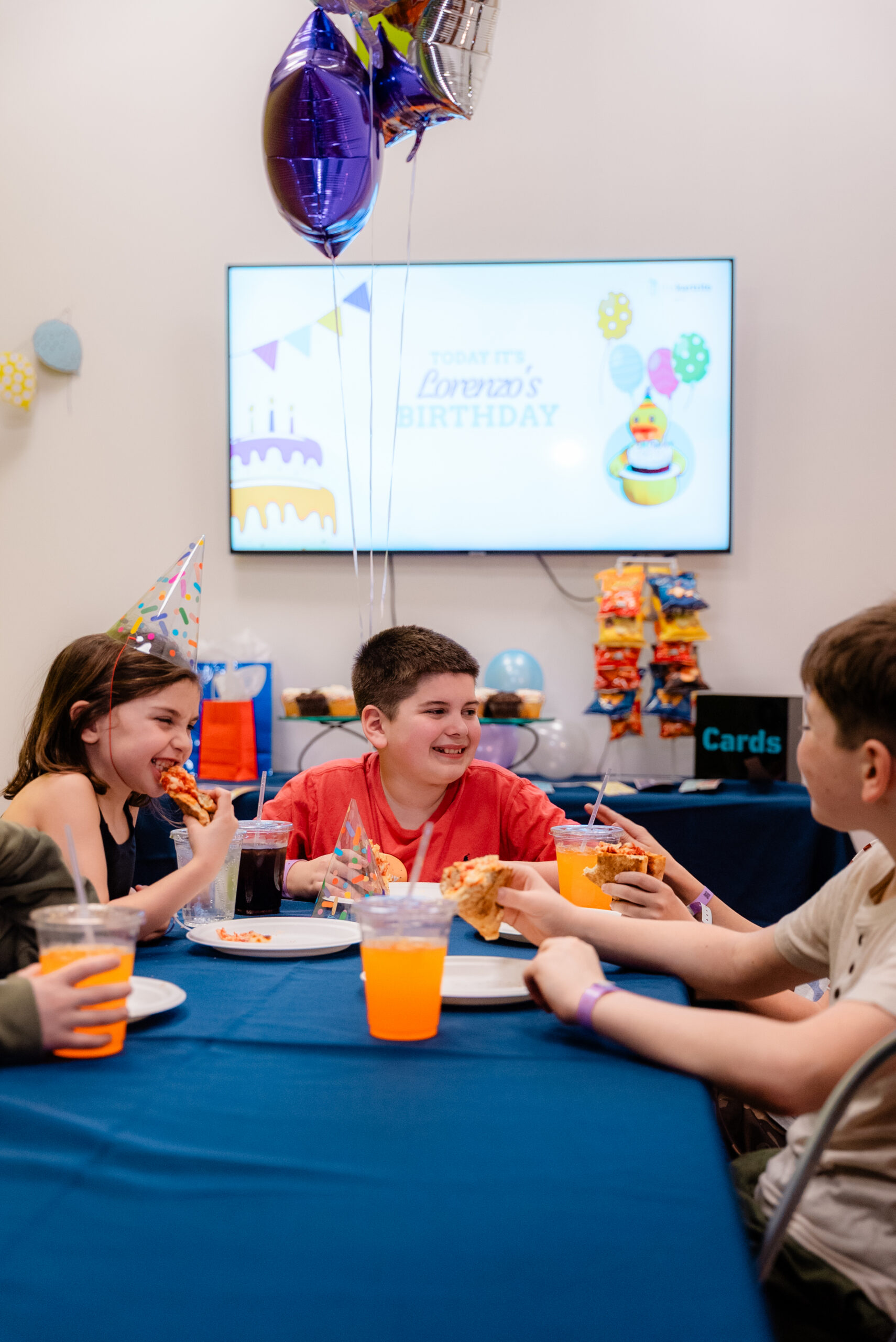 Four kids in party hats eat pizza and drink juice at a birthday party, with balloons and snacks on a decorated table.
