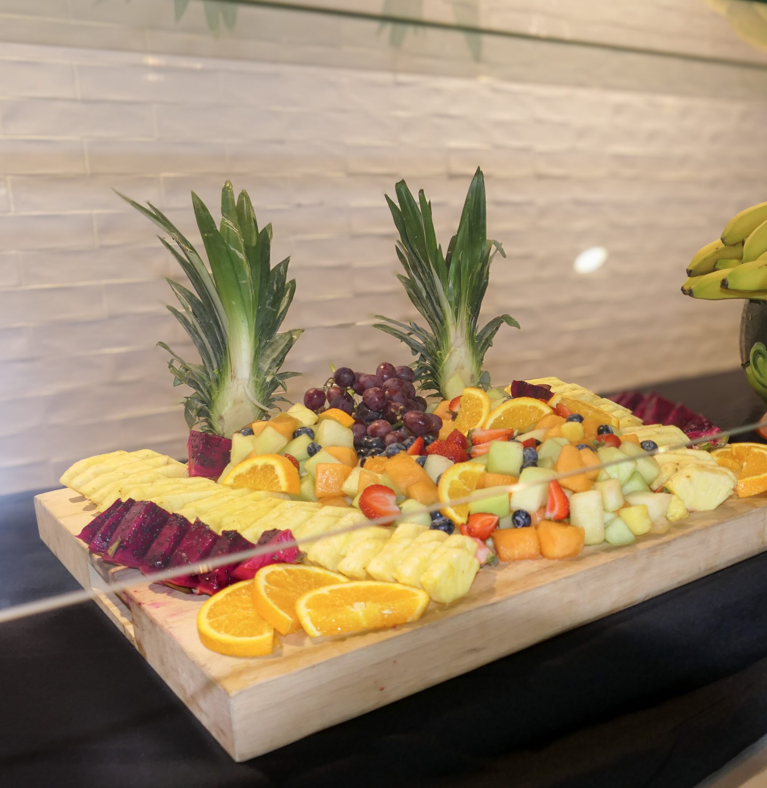 A wooden platter displays sliced pineapples, oranges, dragon fruit, grapes, honeydew, cantaloupe, and blueberries, garnished with pineapple tops.