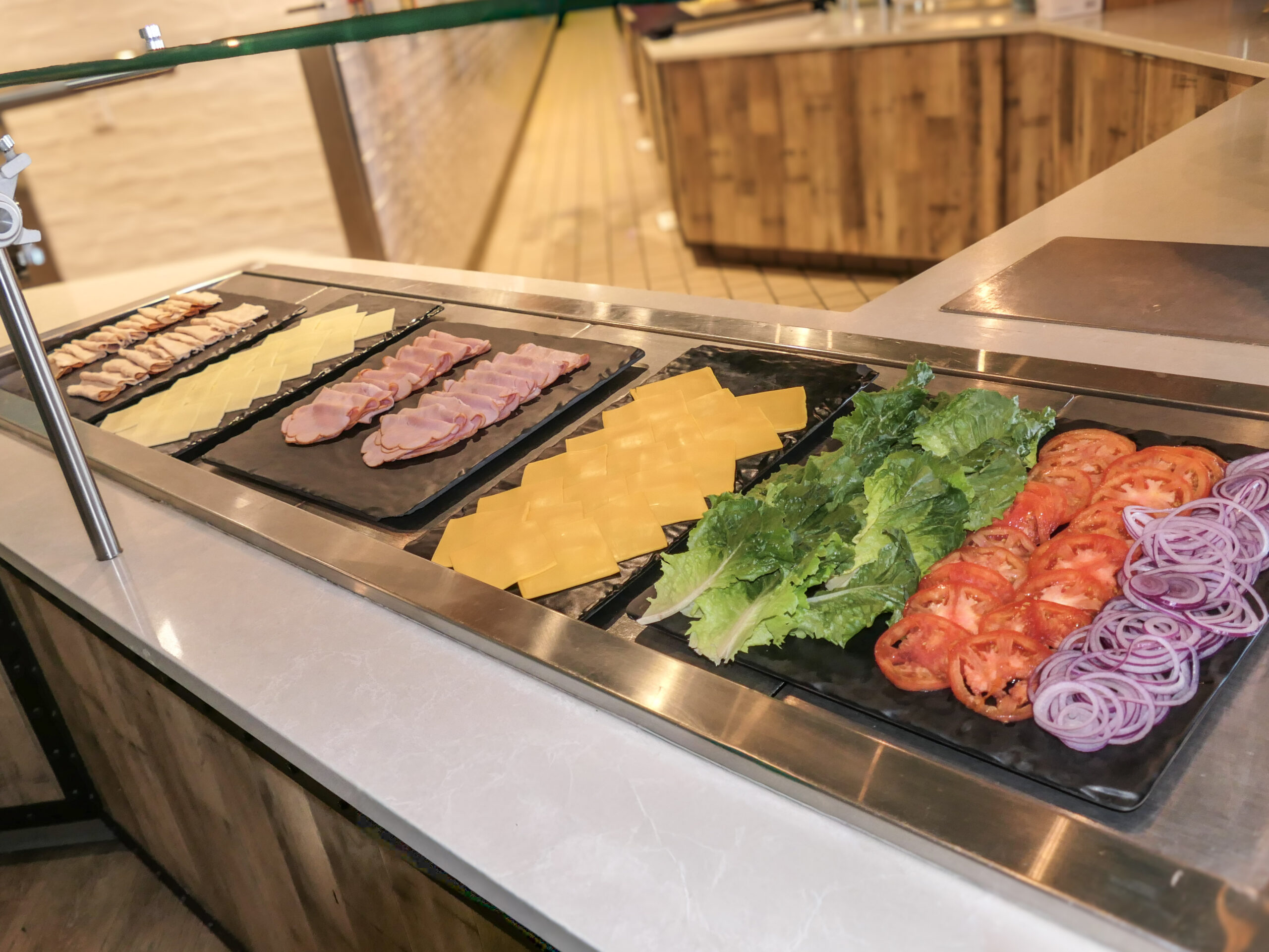 Slices of ham, cheese, lettuce, tomato, and onion are neatly arranged on black trays at a sandwich toppings bar.