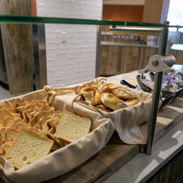 A breakfast buffet with sliced bread, bagels, and assorted condiments in baskets on a wooden counter under a glass partition.