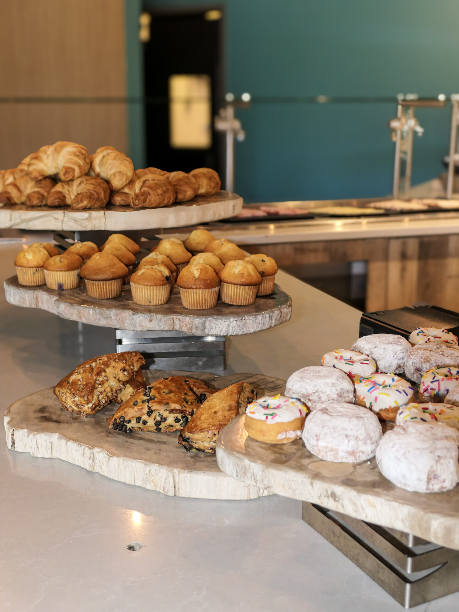 Assorted pastries, muffins, croissants, scones, powdered donuts, and sprinkled donuts arranged on wooden trays in a bakery display.