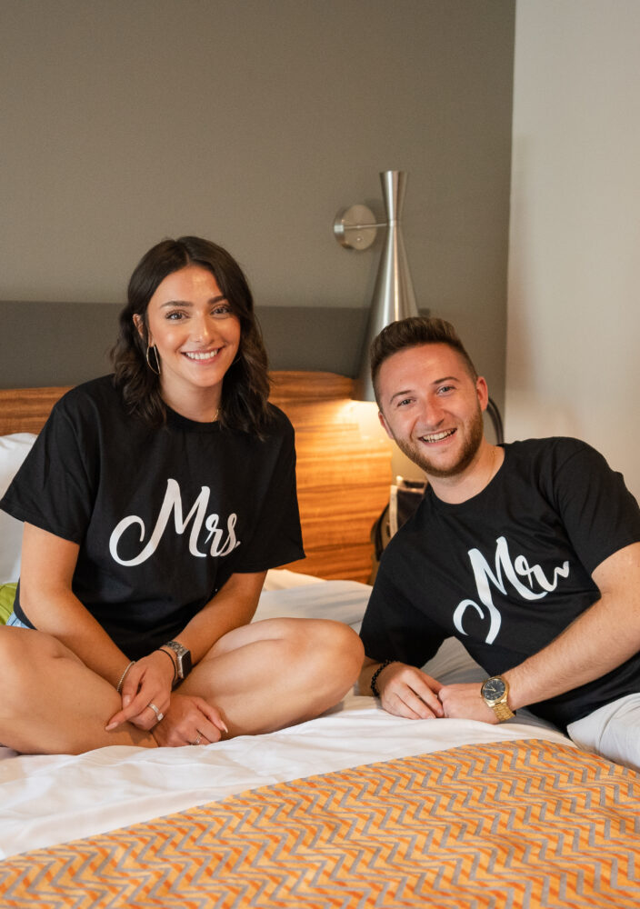 A smiling couple sits on a bed wearing matching black Mr and Mrs shirts, with colorful art on the wall behind them.