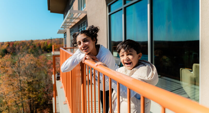 Two children smile and lean over an orange railing on a balcony, with autumn trees and a clear blue sky in the background.