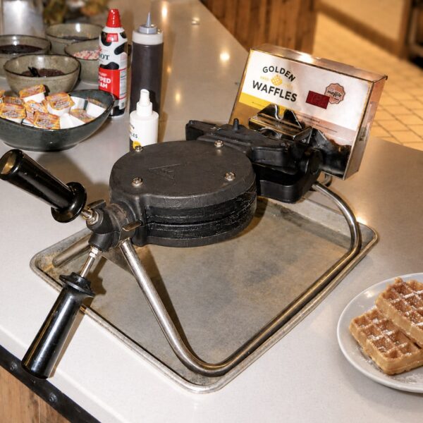 Waffle maker on a counter with batter, syrup bottles, a plate of waffles, and a box labeled Golden Malted Waffles nearby.