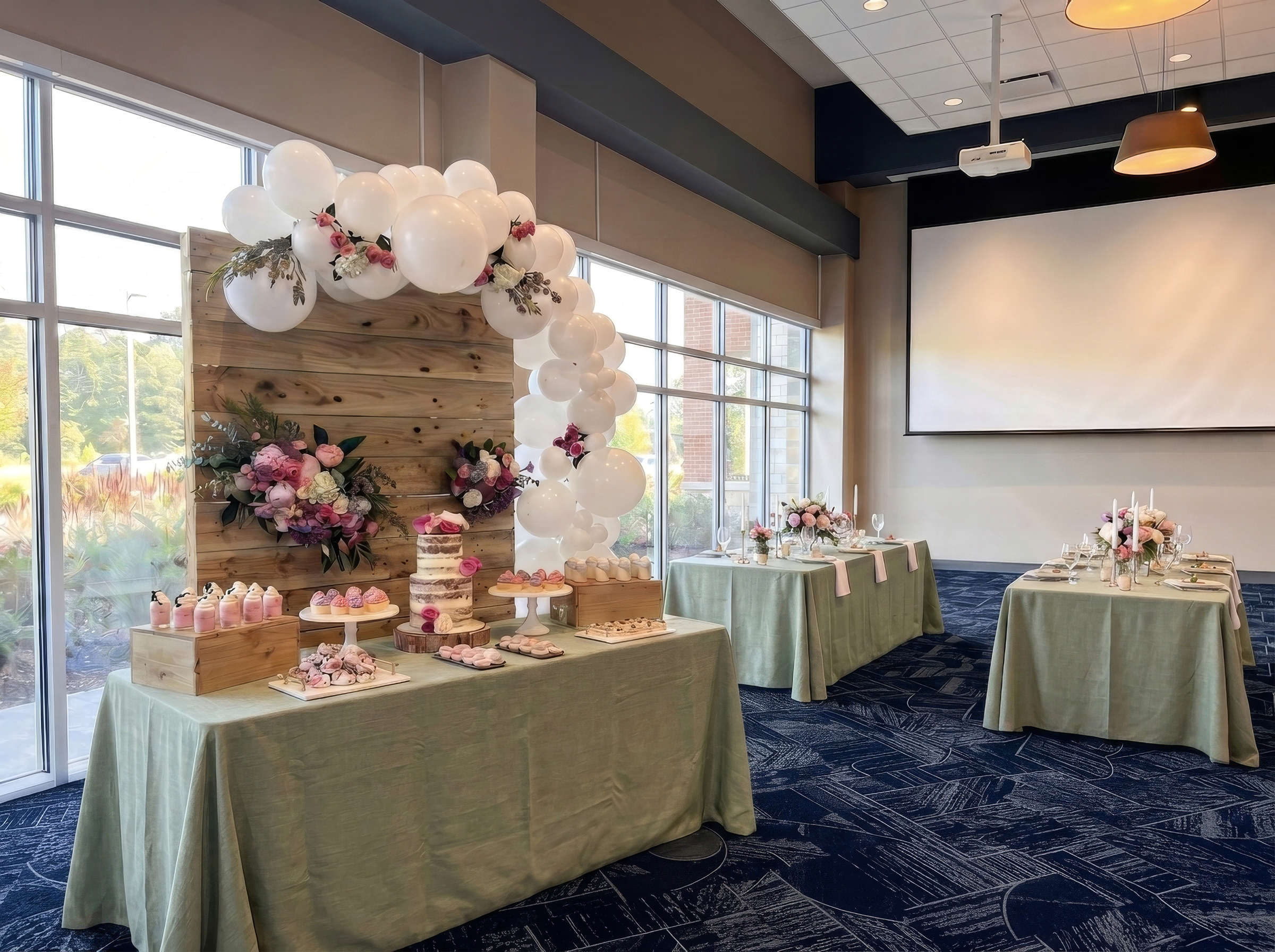 A decorated dessert table with cupcakes and balloons stands in a bright room set up for an event, with tables covered in green tablecloths.