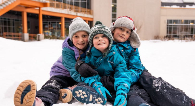 Three children in winter hats and coats huddled together in the snow.