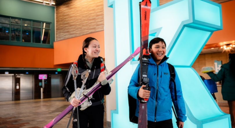 A girl and a boy carrying skis stand in front of a large, glowing blue letter K.